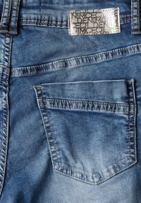 Modrá džínovina s podrobnou zadní kapsou s viditelným šitím a kovovým štítkem s vyraženým textem připevněným na pase.