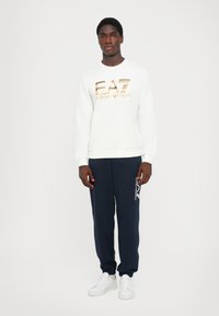 Sweatshirt branco com logo dourado EA7, combinado com calças de treino azul-marinho com um gráfico branco. Os sapatos são brancos e feitos de material liso.