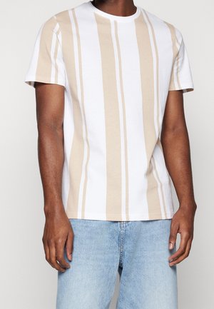 Uomo che indossa una maglietta a maniche corte con larghe strisce verticali beige e bianche e jeans di denim azzurro chiaro.