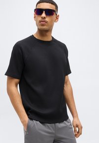 Άνδρας που φοράει μαύρο κοντομάνικο πουκάμισο με υφή, γκρι παντελόνι και μεγάλα αντανακλαστικά γυαλιά ηλίου, με τα χέρια στις τσέπες, μπροστά σε απλό φωτεινό φόντο.