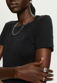 Zwart shirt met korte mouwen en een ronde halslijn, met een zilveren ketting als accessoire. Handen tonen meerdere zilveren ringen en een gestructureerd oppervlak.