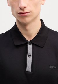 Camiseta polo preta com placket de botões cinza, dois botões e uma textura subtil. Apresenta o logótipo em relevo da BOSS do lado esquerdo.