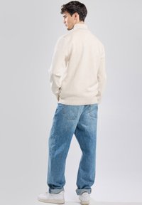 Beige pullover sweater with a high collar, worn with loose-fitting light blue denim jeans and white sneakers. Plain texture, minimal detailing.