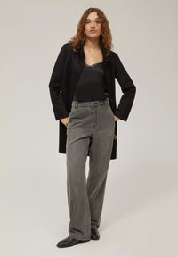 Cappotto lungo nero indossato sopra un top in satin nero, abbinato a pantaloni in denim grigi larghi. Texture lisce e colori in contrasto.