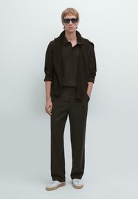 Polo nero sotto un maglione scuro, abbinato a pantaloni larghi scuri e scarpe bianche. Occhiali da sole quadrati completano il look.