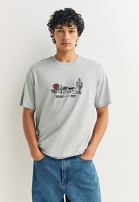 T-shirt à manches courtes gris clair présentant un design graphique avec un soleil, des cactus et le texte "THE BEST OUT IN THE WEST." Coupe décontractée.