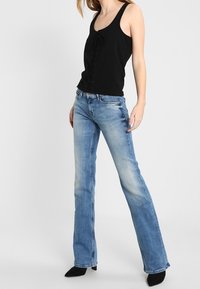 Top negro sin mangas con un detalle de nudo en la parte delantera, combinado con jeans flacos de color azul claro que presentan un desvanecimiento y una textura desgastada, y botas negras de tacón puntiagudo.