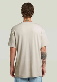 Ljusbeige bomullströja med korta ärmar, rund halsringning och avslappnad passform. Synliga tatueringar på armarna. Enkel, jämn textur.