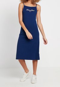 Niebieska bawełniana sukienka midi na cienkich ramiączkach, z haftowanym logo "Tommy Jeans" na piersi i prostą sylwetką. Noszona z białymi sneakersami.