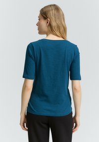Top teal a maniche corte realizzato in un tessuto morbido e testurizzato. Presenta un collo rotondo e una vestibilità rilassata, con un orlo posteriore leggermente più lungo.