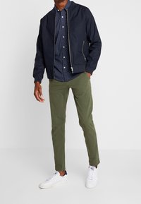 Marine bomberjack met een rits, gecombineerd met een blauwe polka dot overhemd met knopen en olijfgroene broek. Witte sneakers maken de outfit compleet.
