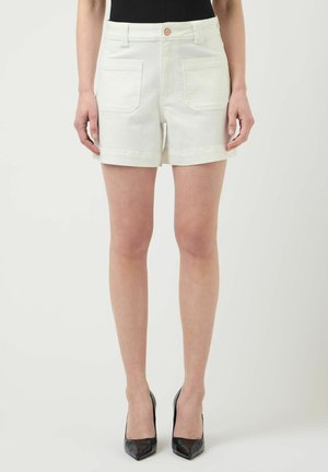 Femme portant un short blanc taille haute avec des poches avant, associé à un haut noir et des talons hauts noirs pointus, debout sur un fond uni.