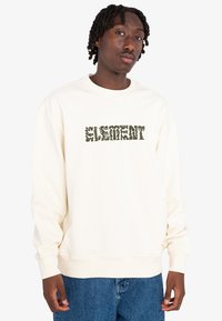 Cream sweatshirt with a textured green "ELEMENT" logo. Features ribbed cuffs and hem, relaxed fit, and soft cotton material. Blue denim jeans worn.