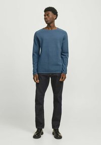 Blå, struktureret sweater med ribbet hals, parret med mørke jeans og sorte sko; modellen står foran en lys baggrund.