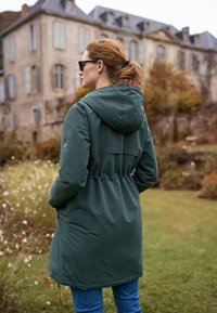 Vrouw met rood haar in een groene jas met capuchon en zonnebril, staand buiten bij een oude stenen gebouw en tuin.