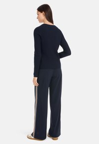 Dunkelblauer gerippter Pullover mit langen Ärmeln, kombiniert mit weiten dunkelblauen Hosen, die über einen gold-weißen Seitenstreifen verfügen, und flachen beigen Schuhen.