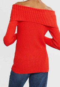 Haut rouge côtelé à épaules dénudées avec des manches longues, présentant un large décolleté. Le matériau présente des lignes texturées verticales. Associé à un bas en denim bleu.