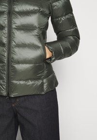 Zelen�á prošívaná bunda s horizontálním švem, se zipem vpředu a bočními kapsami; lesklá, hladká textura; kombinována s tmavými džíny.