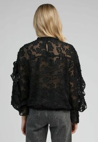 Blouse noire transparente avec broderie florale texturée, coupe décontractée, manches longues, détail en forme de trou de serrure au dos, associée à un jean gris taille haute.