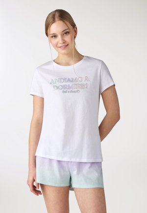 Giovane donna con una maglietta bianca a maniche corte con testo pastello "Andiamo a dormire?" e pantaloncini con sfumatura pastello, sorridente, su sfondo bianco.