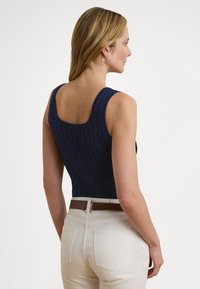 Marinblå stickad linne med kabelmönster, som har en fyrkantig rygg och breda axelband, kombinerad med ljusa jeans och ett brunt bälte.