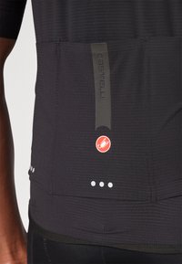 Maillot de cyclisme noir avec un tissu texturé, un panneau gris avec le logo "Castelli", et un logo rouge avec des points d'accent blancs sur le bord inférieur.