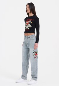 Black long-sleeve cropped shirt with a skull graphic and red text, paired with light blue wide-leg jeans featuring colorful embroidered designs.