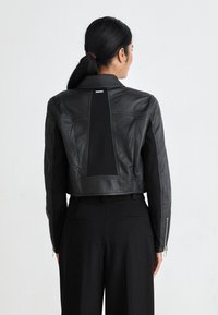 Veste en cuir noir crop avec un design ajusté, présentant un panneau vertical dans le dos et des poignets zippés. Texture lisse et coupe sur mesure.