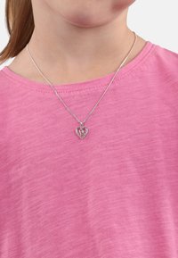 Silberne Halskette mit einem herzförmigen Anhänger, der in einem pinken Oberteil sitzt. Der Anhänger hat filigrane Ausschnitte und kleine Edelsteine, die das Licht reflektieren.