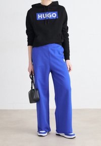 Sudadera negra con el logo "HUGO", combinada con pantalones de chándal sueltos azules. El modelo sostiene un pequeño bolso negro; los zapatos son blancos con acentos azules.