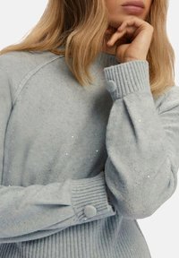Maglione azzurro chiaro in morbido tessuto, con polsini a coste e dettagli in bottone, e una superficie testurizzata con scintillii sottili.