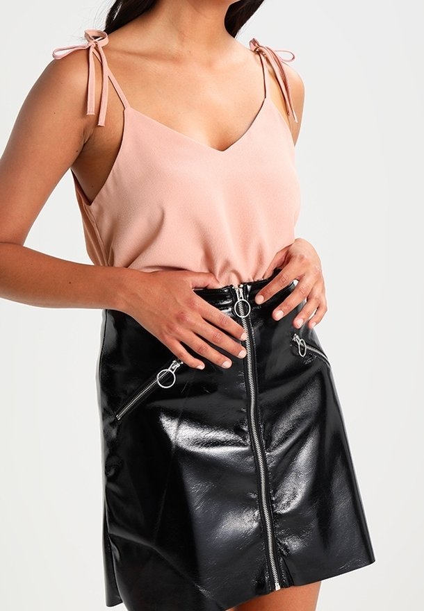 Jasnoróżowa koszulka na cienkich regulowanych ramiączkach w połączeniu z czarną, wysokim stanem spódnicą z ekoskóry, ozdobioną przednim suwakiem i detalami przy kieszeniach.