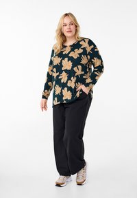 Camicia a maniche lunghe floreale con fiori beige su uno sfondo verde scuro, abbinata a pantaloni neri e sneakers beige con dettagli dorati.