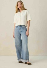 Witte cropped gebreide top met korte mouwen en knoopsluiting, gecombineerd met lichtblauwe wijde jeans met een opgerolde zoom. Zwarte loafers met luipaardprint.