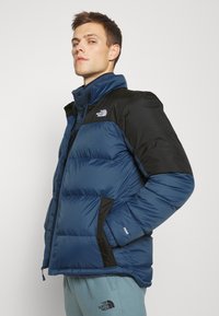 Giubbotto imbottito di colore blu navy con accenti neri. Presenta una chiusura con zip, un alto colletto e una marcata cucitura orizzontale. Design compatto e caldo.