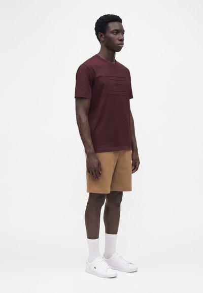 Мъж, облечен с бордова тениска Lacoste, кафяви къси панталони, бели чорапи и бели маратонки на бял фон.