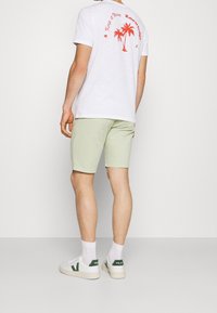 T-shirt bianca in cotone con grafica di una palma rossa sul retro, abbinata a pantaloncini verde chiaro, sneakers bianche con accenti verdi e calzini bianchi.