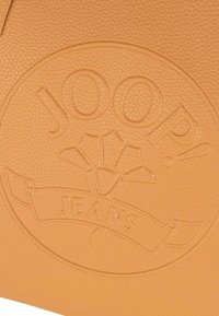 Oranžová kožená taška s vyraženým logem "JOOP! JEANS" s texturovaným povrchem a kruhovým designem, zdůrazňující identitu značky.