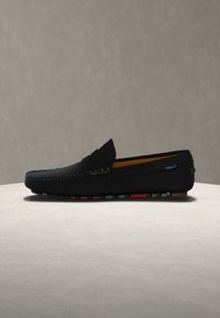 Μαύρο δερμάτινο μοκασίνι με ραμμένες λεπτομέρειες, πολύχρωμη σόλα από καουτσούκ και σχεδιασμό που φοριέται χωρίς κορδόνια, τοποθετημένο σε επίπεδη ανοιχτή επιφάνεια με απλό φόντο.