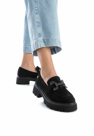 Mocasines de ante negros con suela gruesa y detalle decorativo de lazo. Llevados con pantalones de mezclilla azul claro arremangados.