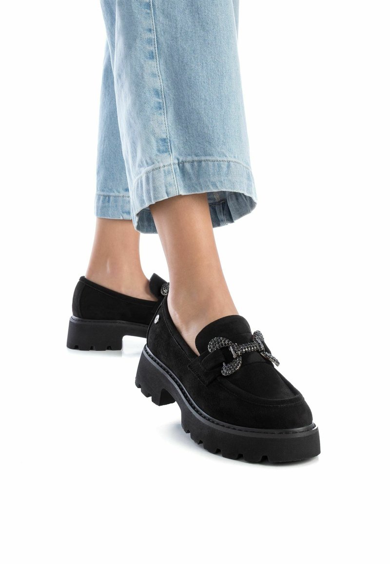 Mocasines de ante negros con suela gruesa y detalle decorativo de lazo. Llevados con pantalones de mezclilla azul claro arremangados.