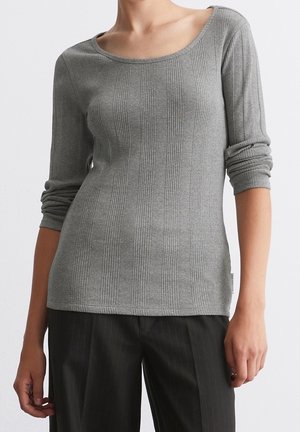 Frau trägt ein eng anliegendes graues, geripptes Langarmshirt mit gerafften Ärmeln und schwarze, schwarz gestreifte Hose, steht vor einem schlichten weißen Hintergrund.