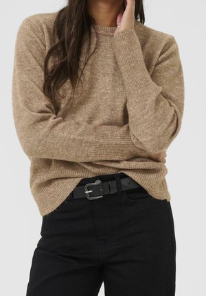 Vrouw draagt een beige gebreide trui met lange mouwen, zwarte broek en een zwarte riem met een zilveren gesp, hand aan haar gezicht.