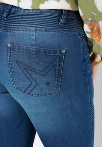 Jeans de denim azul con un bolsillo trasero que presenta un detalle de costura en forma de estrella y zigzag. Los jeans tienen una cinturilla texturizada y un ajuste clásico.