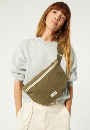 Olivgrüner Canvas-Umhängetasche mit einem weißen Reißverschluss, ausgestattet mit einem frontalen Patch-Label. Kombiniert mit einem grauen Sweatshirt und cremefarbenen Hosen.