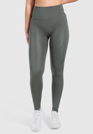 Grüne Leggings aus dehnbarem Material mit hoher Taille, seitlichen Taschen und kontrastierenden Nähten, kombiniert mit weißen Sportschuhen.