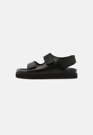 Sandalia negra con correas ajustables de velcro, diseño abierto, suela plana y textura suave. Cuenta con recortes en la parte superior para mayor transpirabilidad.