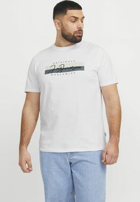 Bílý bavlněný tričko s grafickým designem v černé a šedé barvě a textem "JACK & JONES", kombinované se světle modrými džíny.