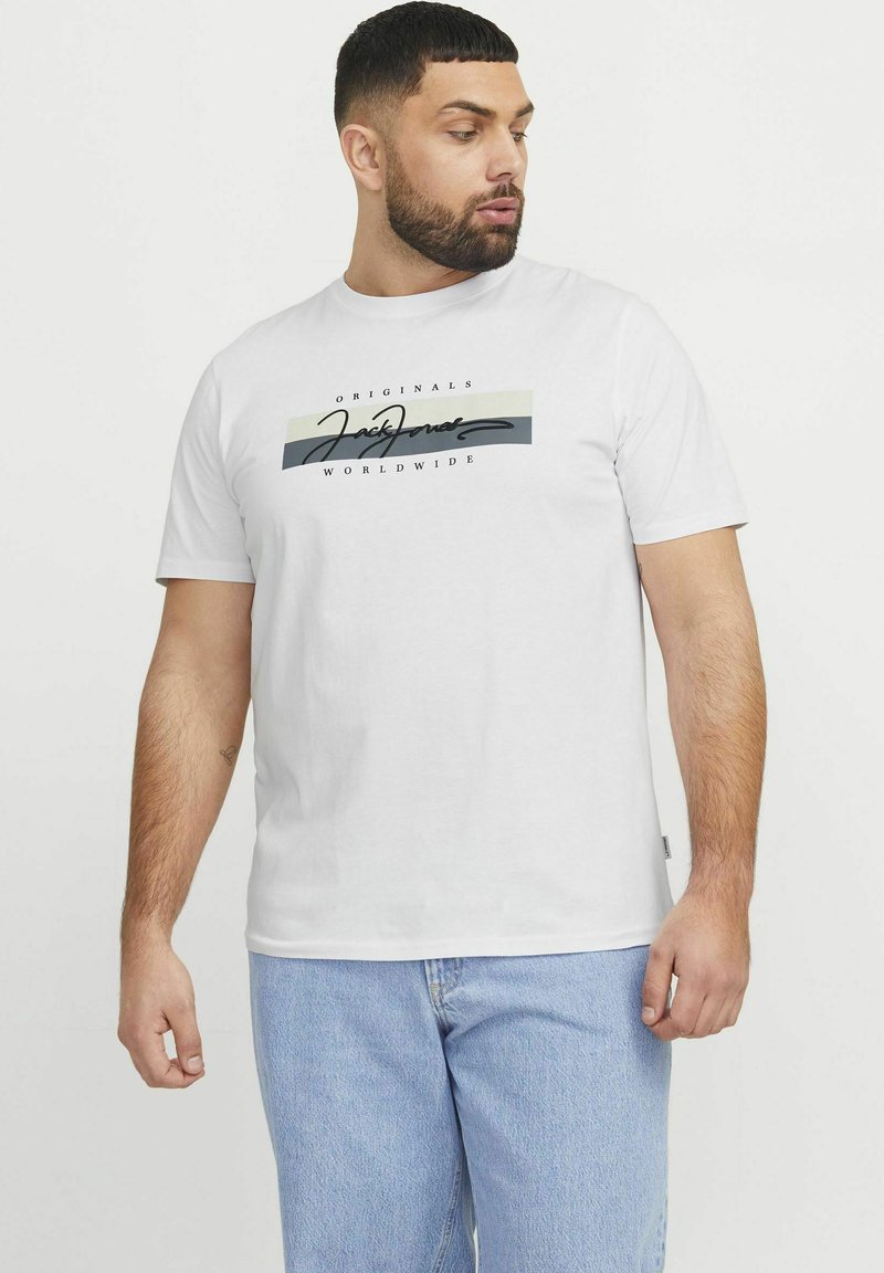 Balts kokvilnas T-krekls ar grafisko dizainu melnos un pelēkos toņos un tekstu "JACK & JONES", apvienots ar gaiši ziliem džinsu biksēm.