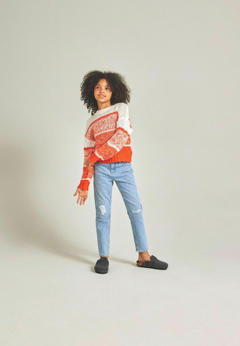 Maglione lavorato a maglia arancione e bianco con righe orizzontali, abbinato a jeans strappati blu chiaro e scarpe slip-on grigio scuro.